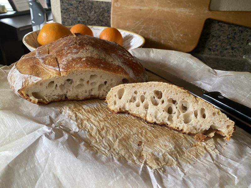 A golden domed loaf on parchment with one thick slice cut away, the slice showing a holey open crumb, a bread knife and a bowl of oranges behind it