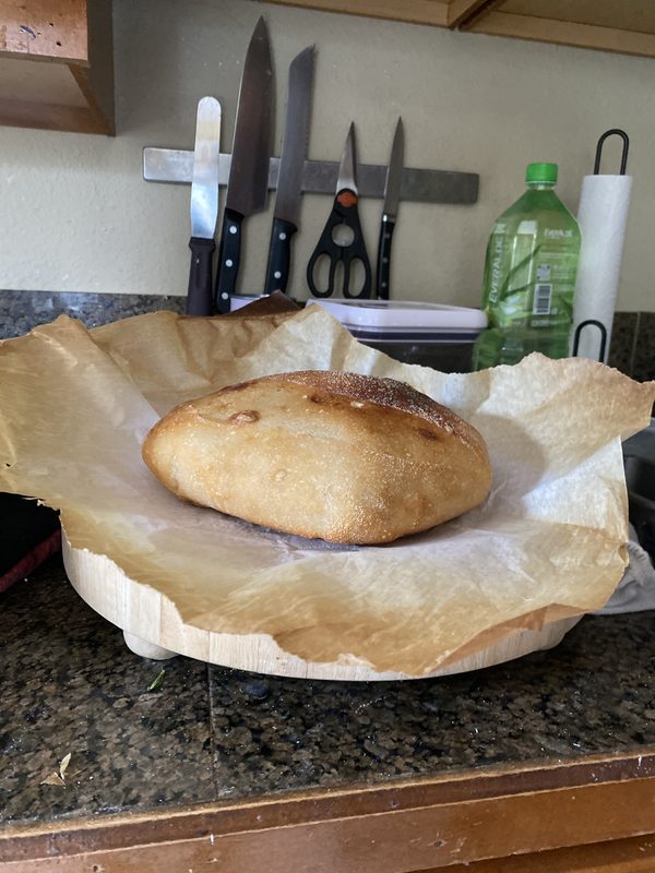 A low, wide golden-brown oval loaf sitting on parchment on a round wooden board on a kitchen counter