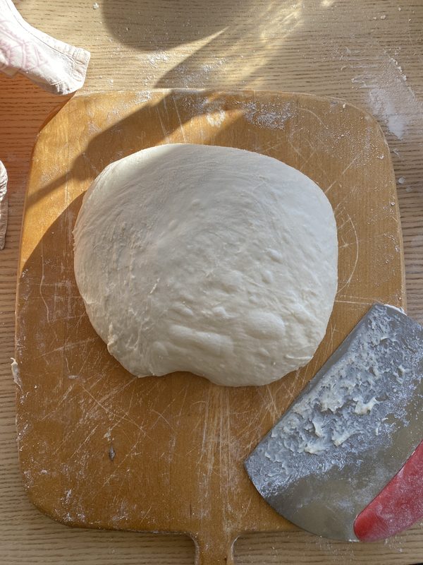 A shaped round of raw white dough resting on a floured wooden peel in sunlight, with a metal bench scraper beside it